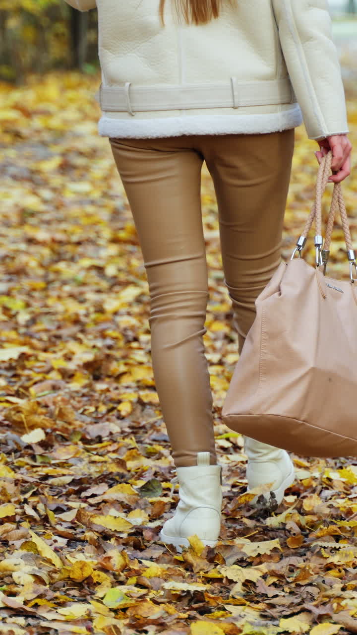 Female legs in white boots walk by the colorful carpet of leaves in the park. Unrecognized woman with big bag followed by camera. Vertical video
