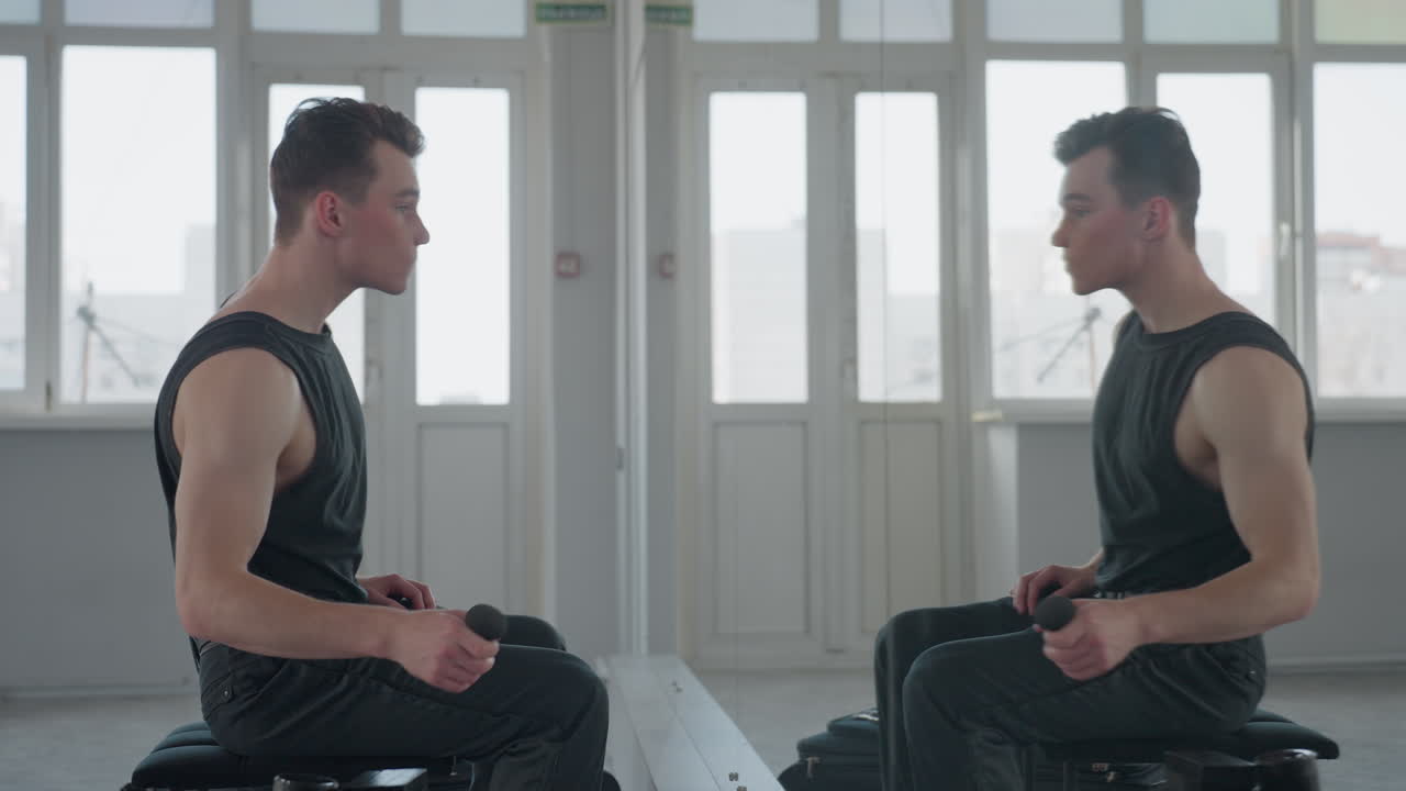 man in black vest sits before large mirror in bright studio, holds makeup applicator near face, gently applies product while observing reflection, urban buildings visible through tall windows
