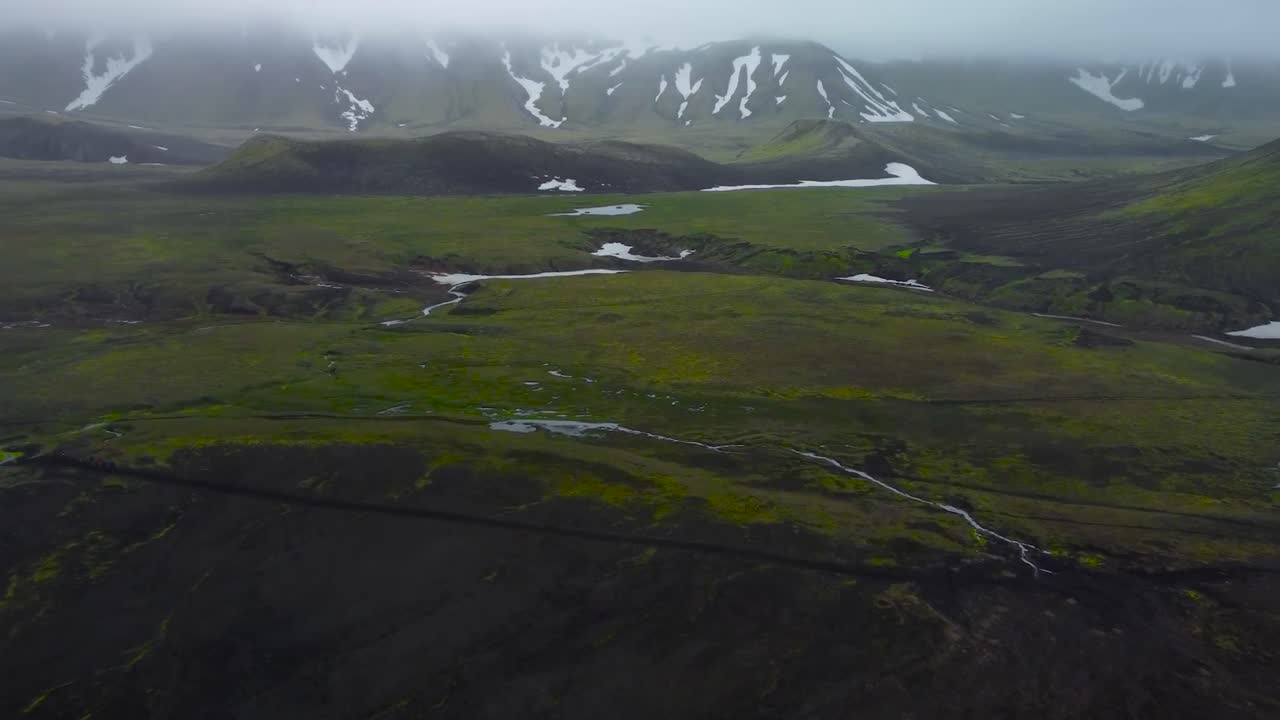 Aerial drone view of green mossy and dark rocky terrain in Iceland with snow covered mountains in the background. A small narrow river is flowing on the volanic dark ground between mossy snowy terrain