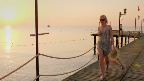 a carefree woman in a light dress with a hat in her hand walks along the pier