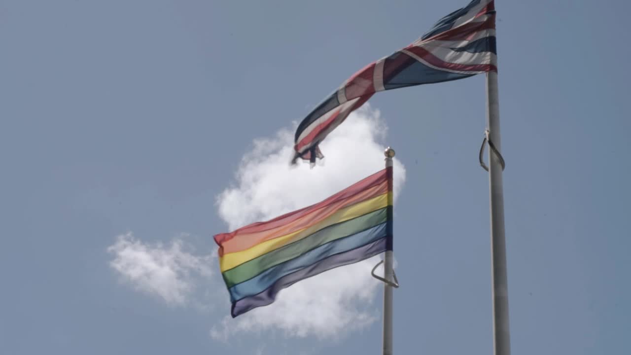 Union Jack and Rainbow Pride flag flying against blue sky and clouds wide shot