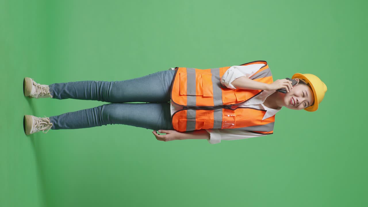 안전 헬을 입은 아시아 여성 엔지니어의 몸 전체가 녹색 화면 배경 스튜디오에 서서 스마트폰으로 이야기하고 있습니다.