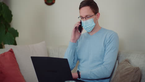 Man-Wearing-Protective-Mask-While-Working-On-Laptop-And-Talking-On-Mobile-Phone