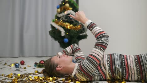Feliz-Chica-Festiva-Tirada-En-El-Suelo-Con-Confeti-Dorado-Brillante-Y-Tomándose-Selfie.-árbol-De-Navidad-Decorado-En-El-Fondo