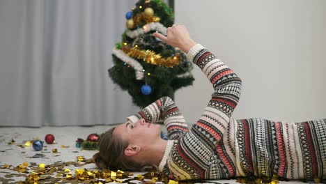 feliz chica festiva tirada en el suelo con confeti dorado brillante y tomándose selfie. árbol de navidad decorado en el fondo