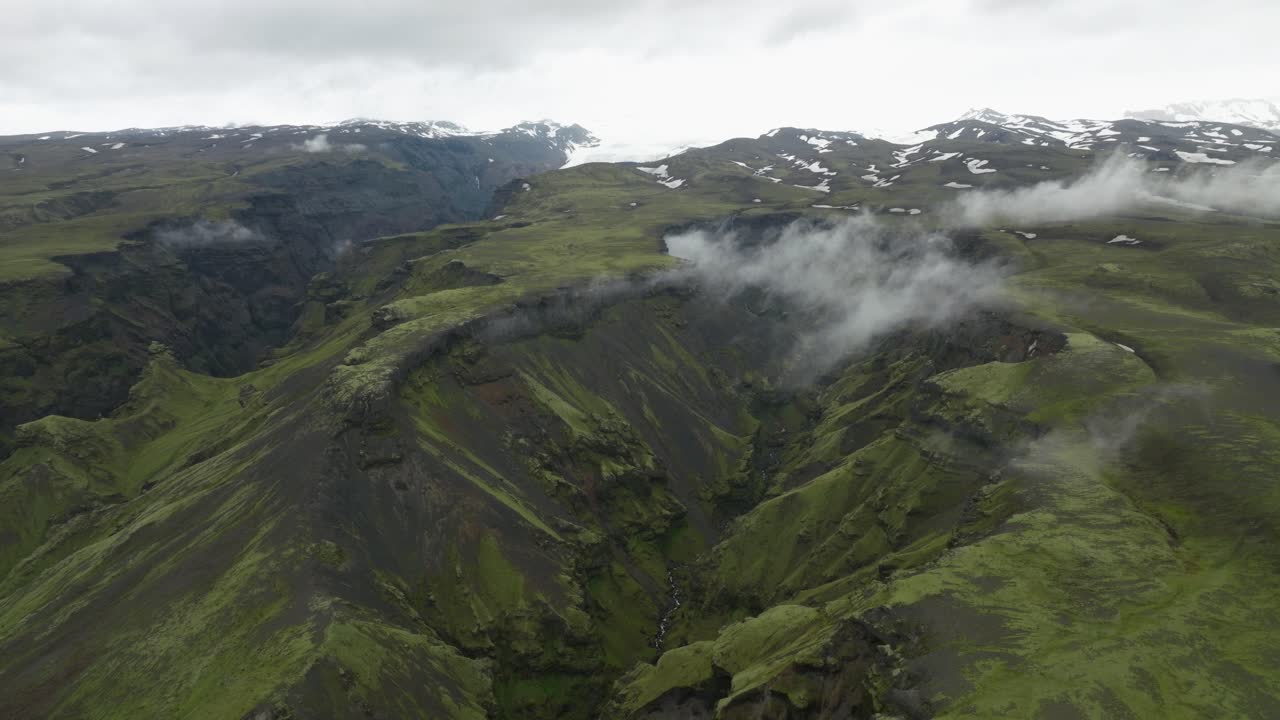 An expansive aerial view of a lush green valley and rugged cliffs in Hafursey, Iceland, with patches of mist and distant glaciers.