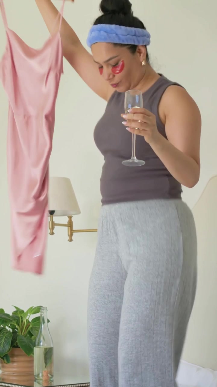 Video verticale: Donna con fascia per capelli che si china per scegliere un vestito in camera da letto, tenendo un bicchiere e prendendo i tacchi.