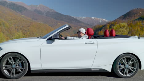 man driving a white convertible through scenic mountains in autumn