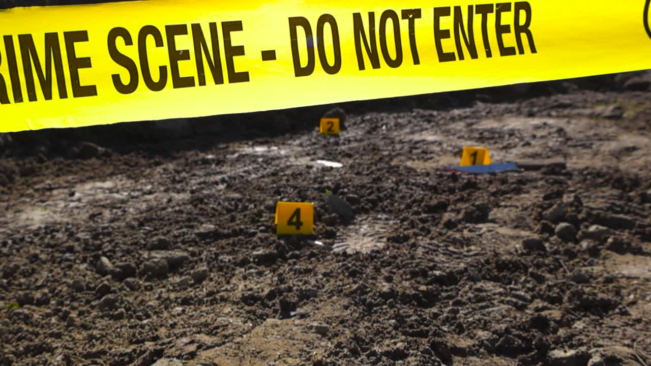 Yellow police line crime scene forensic investigation tape ribbon in front of crime scene police markers next to bloody knife, footprint and drugs on a muddy road in sunny rural countryside