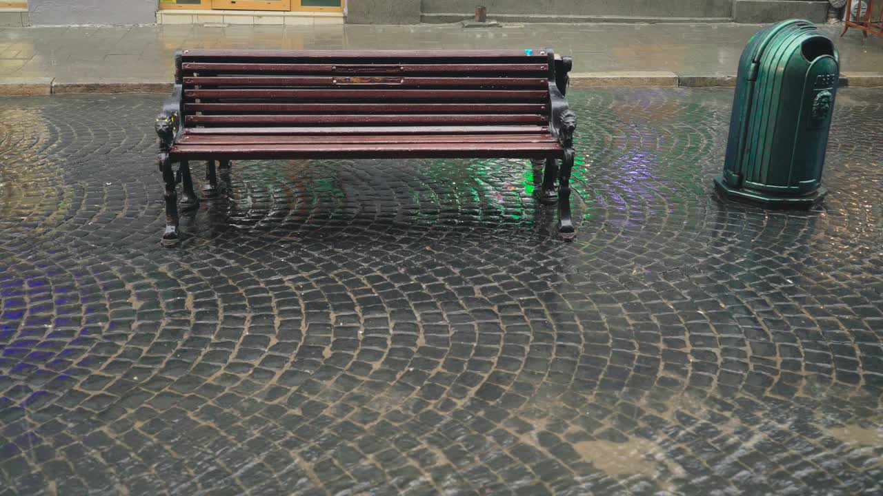 Heavy rain is falling on the pavement and modern wooden bench outdoors in the city. Stream of water are flowing through the pavement in a rainy weather.