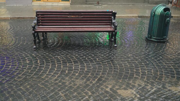 Heavy rain is falling on the pavement and modern wooden bench outdoors in the city. Stream of water are flowing through the pavement in a rainy weather.