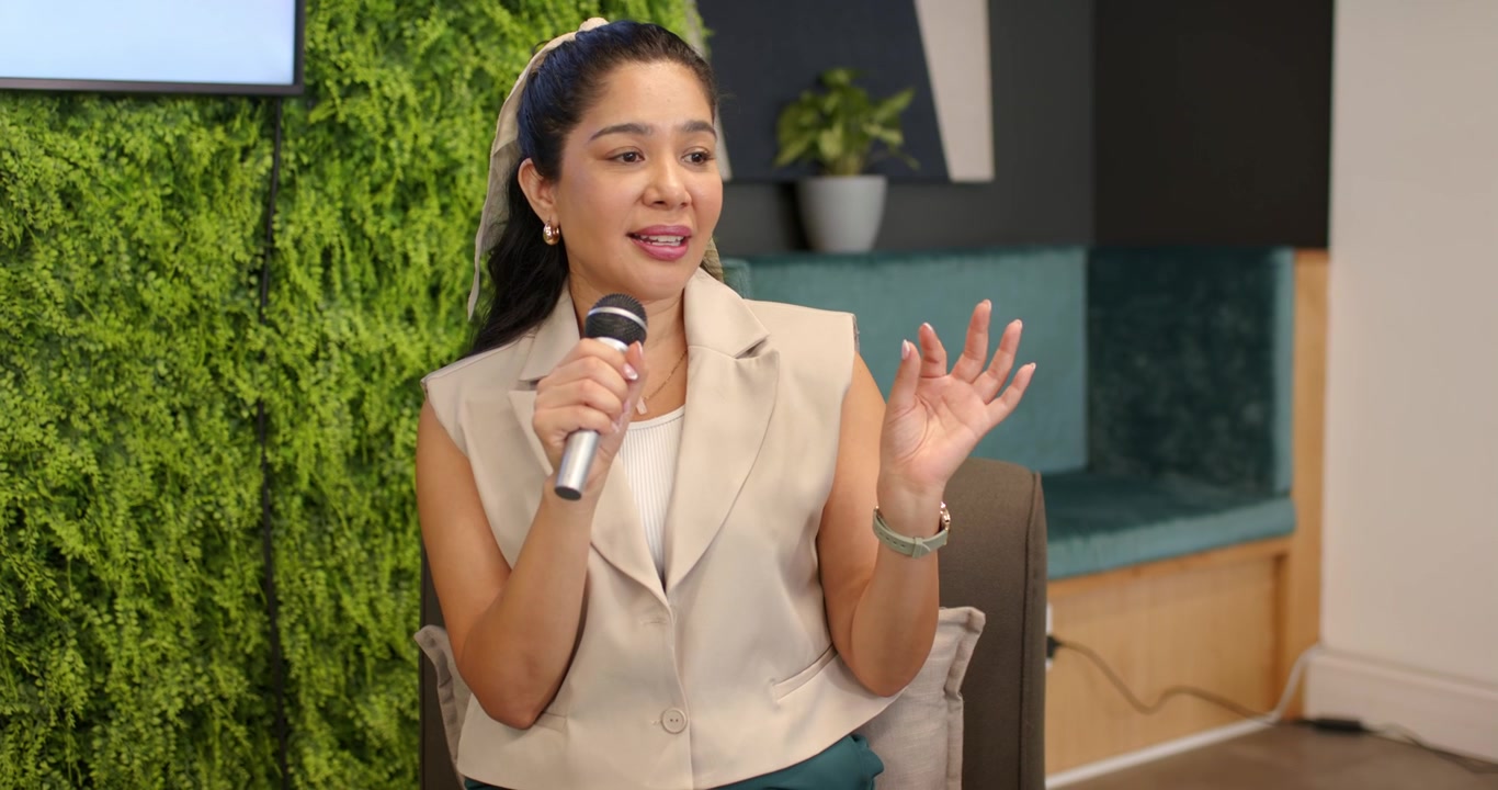 Adult female speaker holding mic and gesturing at modern lounge while slide causing brief pause
