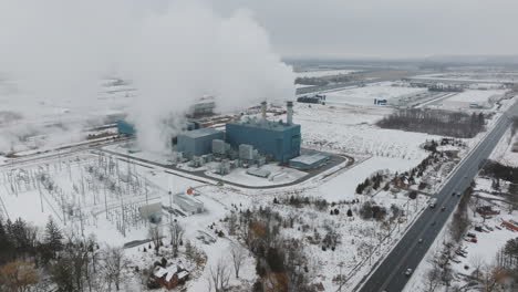 Central-Eléctrica-Industrial-Que-Emite-Vapor-En-Un-Paisaje-Nevado,-Autopista-En-Primer-Plano,-Vista-Aérea