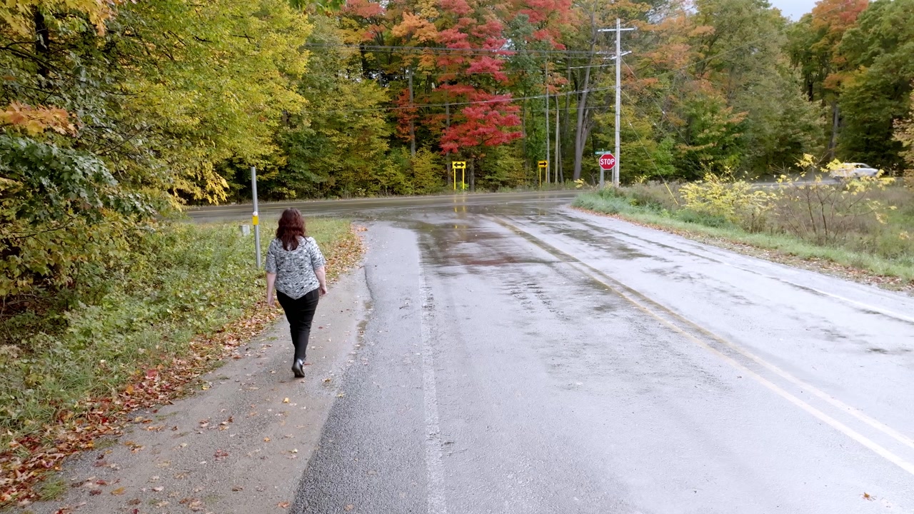 Middle aged caucasian woman walking along a road in Northern Michigan with fall colors and drone video following.