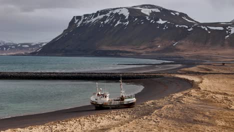 aerial drone video circling an abandoned shipwreck beached on a black sand beach on a cloudy day in iceland with snowcapped mountains off in the distance