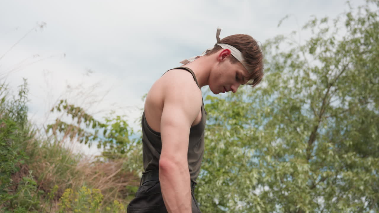 white man standing on grassy slope brushing dust from cargo pants pocket revealing phone outline while surveying surroundings after scramble up bank under bright cloudy sky in relaxed enviroment