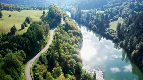 winding country road along bright blue river in which the bright sun is reflected surrounded by green forests, aerial drone