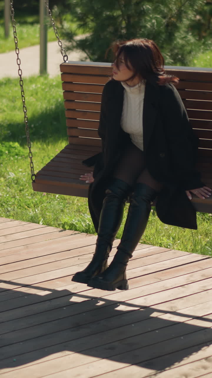 joven con elegante abrigo negro y botas se sienta en un columpio de madera en un parque iluminado por el sol, mirando serenamente a la distancia, rodeada de exuberante vegetación y un jarrón de flores blancas