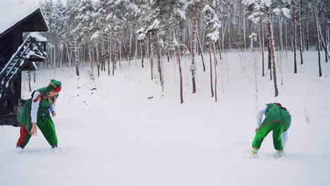 two elves in green clothing are playing snowballs together on the forest background. happy elves male and a female are having a snowball fight outdoors near the wooden house. slow motion.