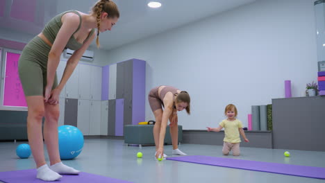 women in gym interacting with baby crawling on floor while one woman in brown offers tennis ball to mother, background features workout equipment, yoga mats, and gym decor