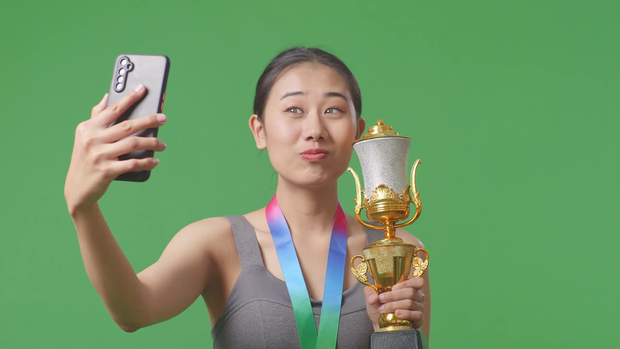 Close Up Of Asian Woman With A Gold Medal Holding A Gold Trophy And Taking Photo On Smartphone As The First Winner On Green Screen Background In The Studio