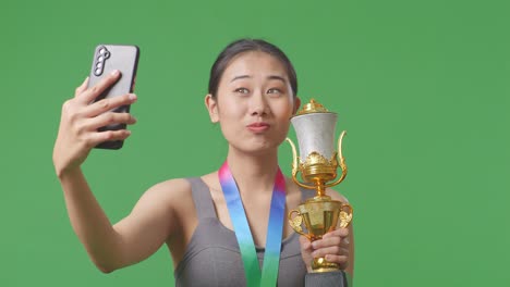close up of asian woman with a gold medal holding a gold trophy and taking photo on smartphone as the first winner on green screen background in the studio