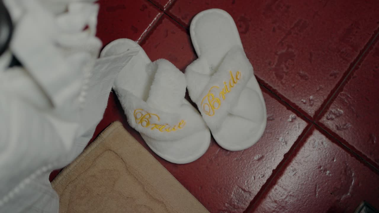 White slippers with 'Bride' in gold embroidery on a red tile floor with a beige mat nearby
