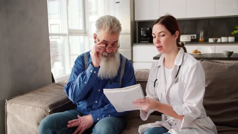 un hombre mayor con cabello gris y una barba exuberante se pone gafas para leer lo que una mujer morena médico en una bata blanca quiere transmitir a él durante un examen en casa en el sofá en un apartamento moderno