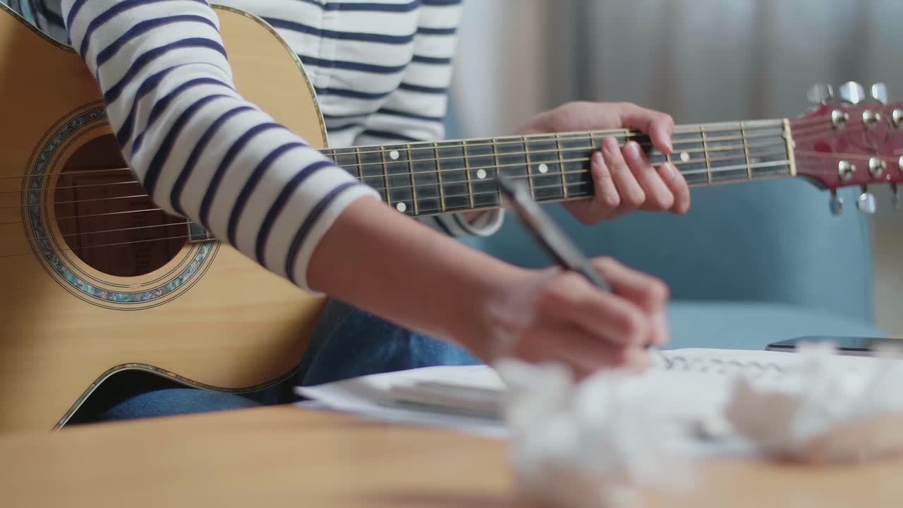 primer plano de una mujer asiática compositora con una guitarra papel arrugado y tirarlo antes de tener dolor de cabeza mientras compone música en casa