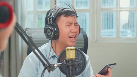 over shoulder view, young asian boy holding phone and wearing headphones singing into microphone while recording podcast with kid girl host in studio
