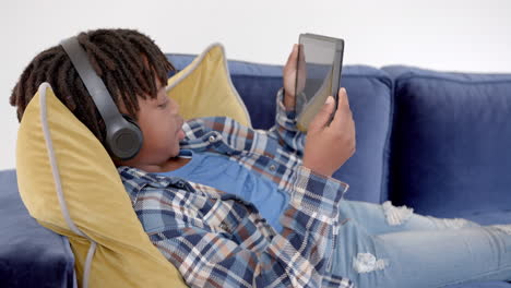 african american boy with headphones using tablet lying on sofa at home, slow motion