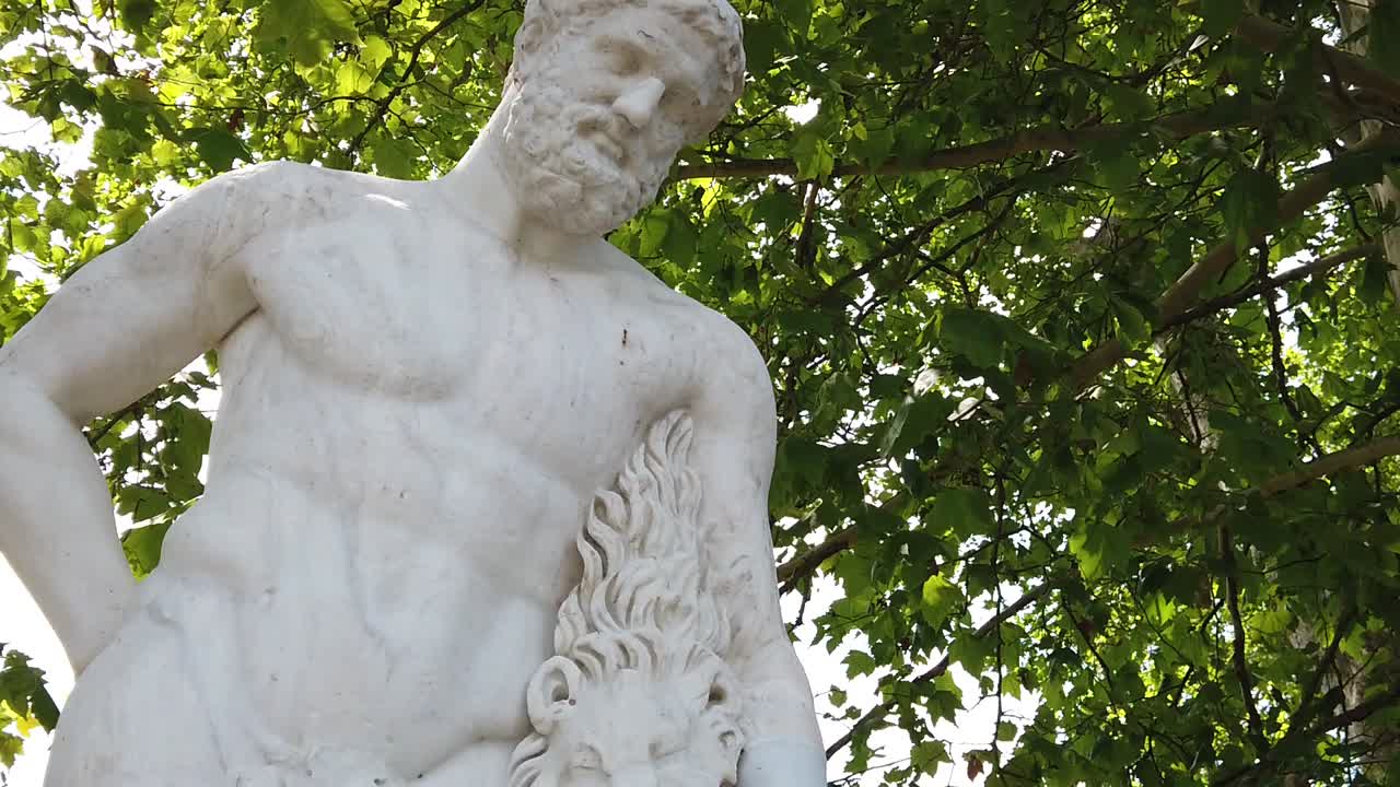 Primo piano dinamico di una classica statua greca in marmo di Ercole che tiene una pelle di leone, con luce solare filtrata attraverso un lussureggiante fogliame verde, texture lisce, composizione artistica, senza persone.