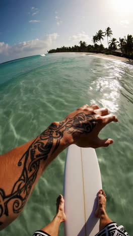first-person view of a surfer holding a board in clear tropical water with a beach and palm trees in the background.
