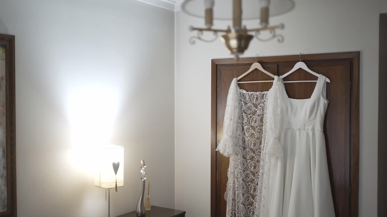Two white wedding dresses hanging on wooden door inside room with lamp and chandelier visible in interior