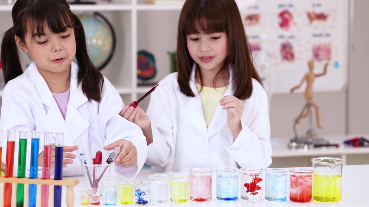 白衣を着た2人の少女が、スポイトとビーカーを使って色鮮やかな化学実験を行っている
