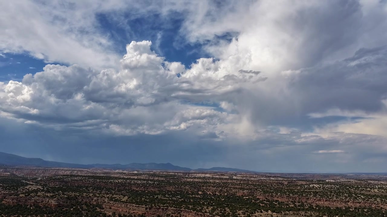 4K drone-timelapse som stiger over en vidstrakt ørkenmesa. Raskt bevegelige, turbulente hvite stormskyer og mørkegrå værsystemer beveger seg raskt over en vid blå himmel over steinete terreng og mørk kratt.