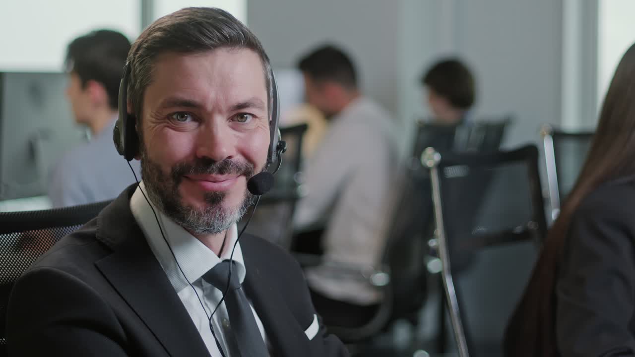 Portræt af en glad og smilende mand, der kigger ind i kameraet, mens han arbejder i et callcenter. Han bruger en stationær pc ved et computerbord og en telefonlinje i et moderne co-working-miljø.