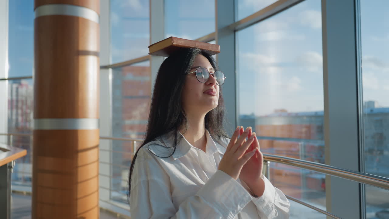 Woman with glasses carefully walking while balancing book on head inside modern glass building, dressed in white shirt and black pants, sunlight streaming through large windows with urban view