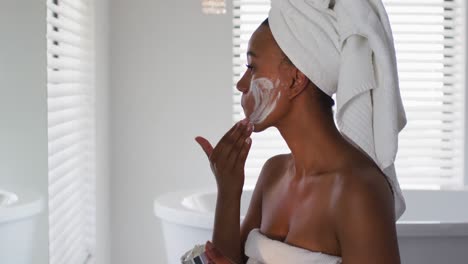 Afroamerikanische-Frau,-Die-Gesichtsmaske-Aufträgt-Und-In-Den-Spiegel-Im-Badezimmer-Schaut