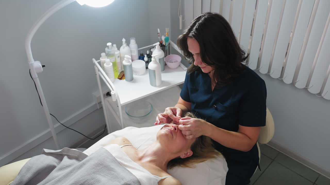 High angle view showing skin care expert using gentle circular strokes from forehead toward eyebrow on relaxed client lying on massage bed under soft clinic light facial rejuvenation treatment