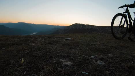 Chica-Empujando-Su-Bicicleta-En-Una-Colina-De-Montaña-Al-Atardecer-Con-Un-Hermoso-Lago-De-Cañón-En-El-Fondo