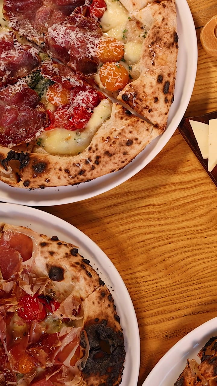 Una telecamera dall'alto riprende pizze gourmet, salumi e formaggi su un tavolo rustico di un ristorante
