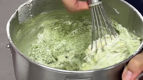 hand mixing flour, sugar, butter, matcha powder in a mixing bowl with a whisk, close up shot