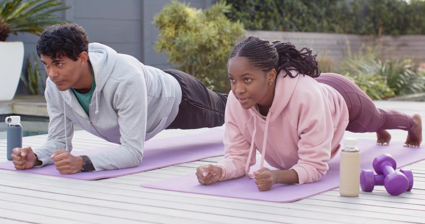 Diverse workout partners holding forearm plank on patio deck following timed core set with mats