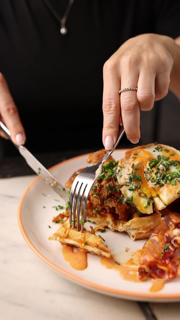 Nahaufnahme von Händen, die Hähnchen und Waffeln mit Spiegelei schneiden