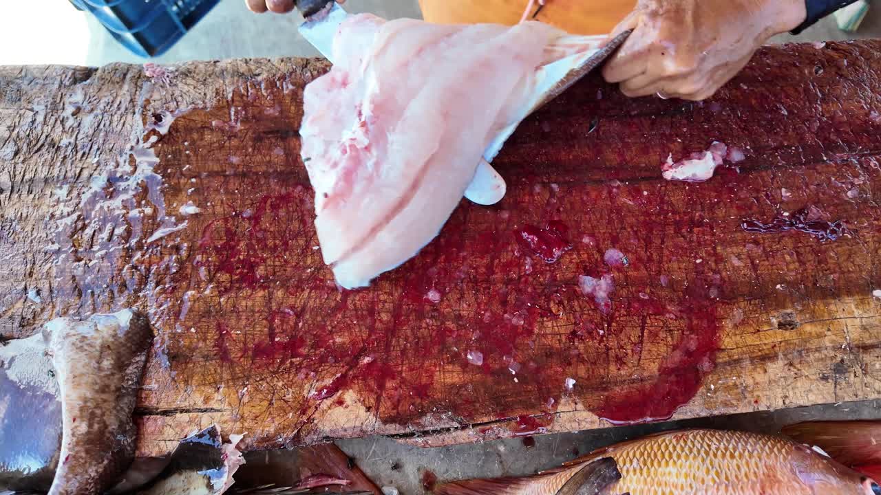 Primo piano di pesce fresco sfilettato a mano su un tagliere di legno rustico, con l'azione visibile del coltello e la consistenza cruda. Dettagli organici, industria della pesca, gastronomia e processi culinari autentici.