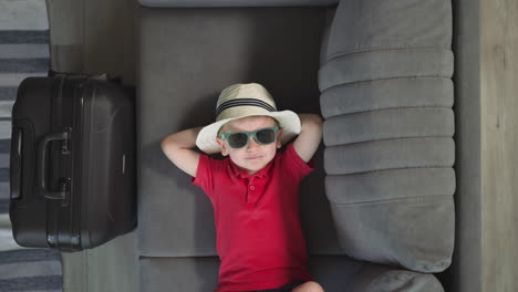 tired little boy in sunglasses lies on sofa near suitcase