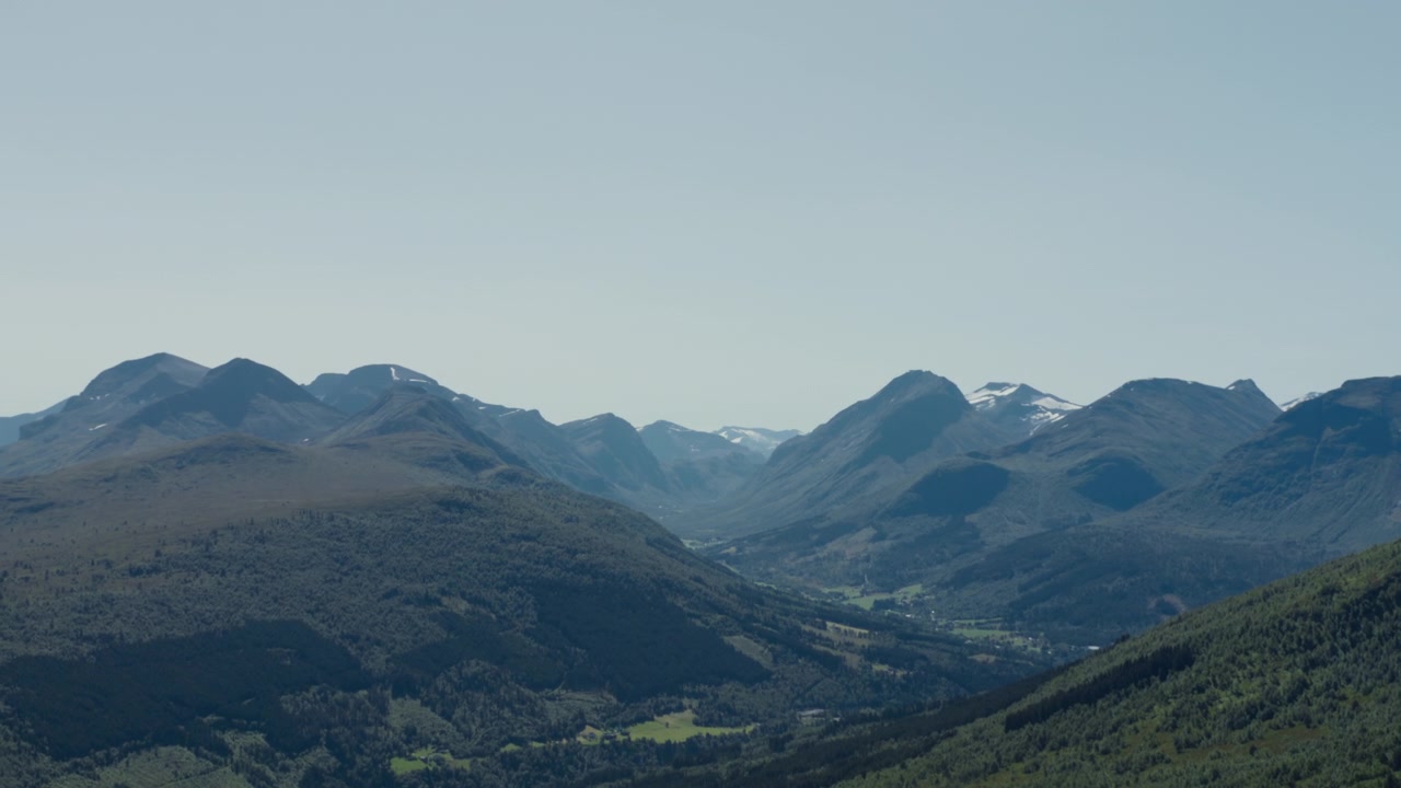 Pegunungan fjell berhutan berlapis mengelilingi lembah hijau lurus di bawah langit biru pucat di Sunnmøre, Norwegia