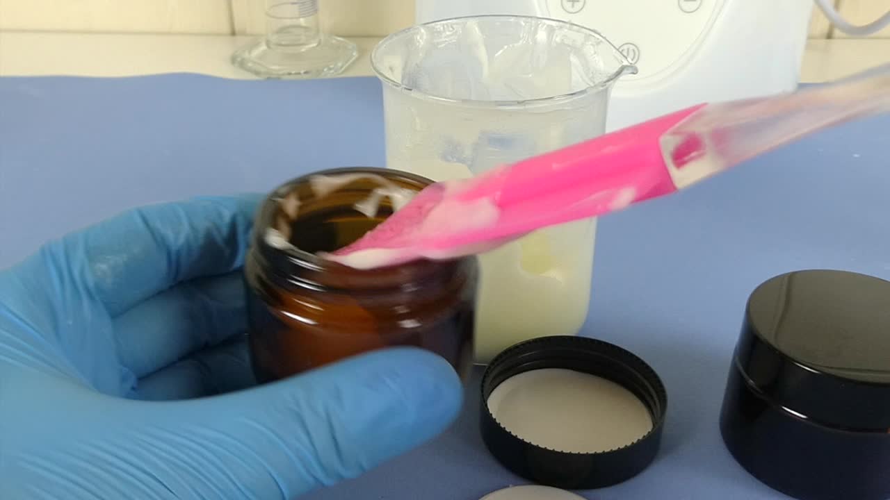 Macro view of a cosmetic laboratory scene where a gloved operator uses a spatula to carefully package a white emulsion.
