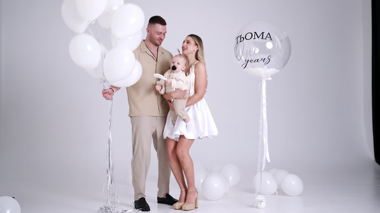 Caucasian family celebrating the first birthday of their baby. Happy parents dance with the child and balloons in studio.